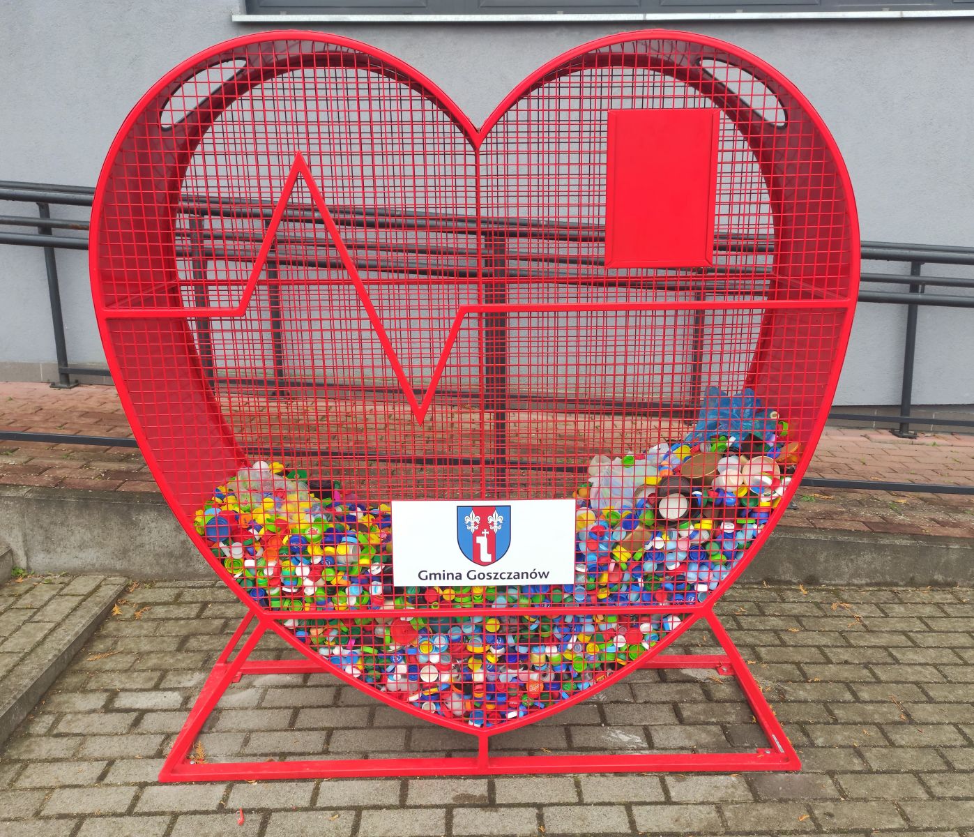 zdjęcie przedstawiajace czerwony pojemnik w kształcie serca na nakrętki z logo Gminy Goszczanów